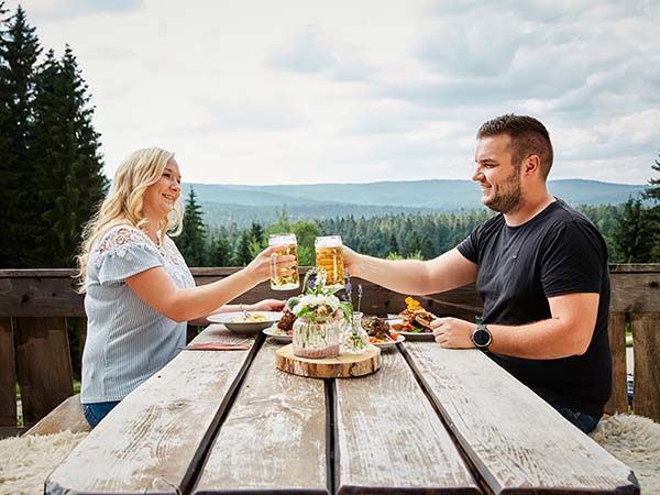 Paar sitzt auf Terrasse eines Gasthauses und stößt mit zwei Bierkrügen an