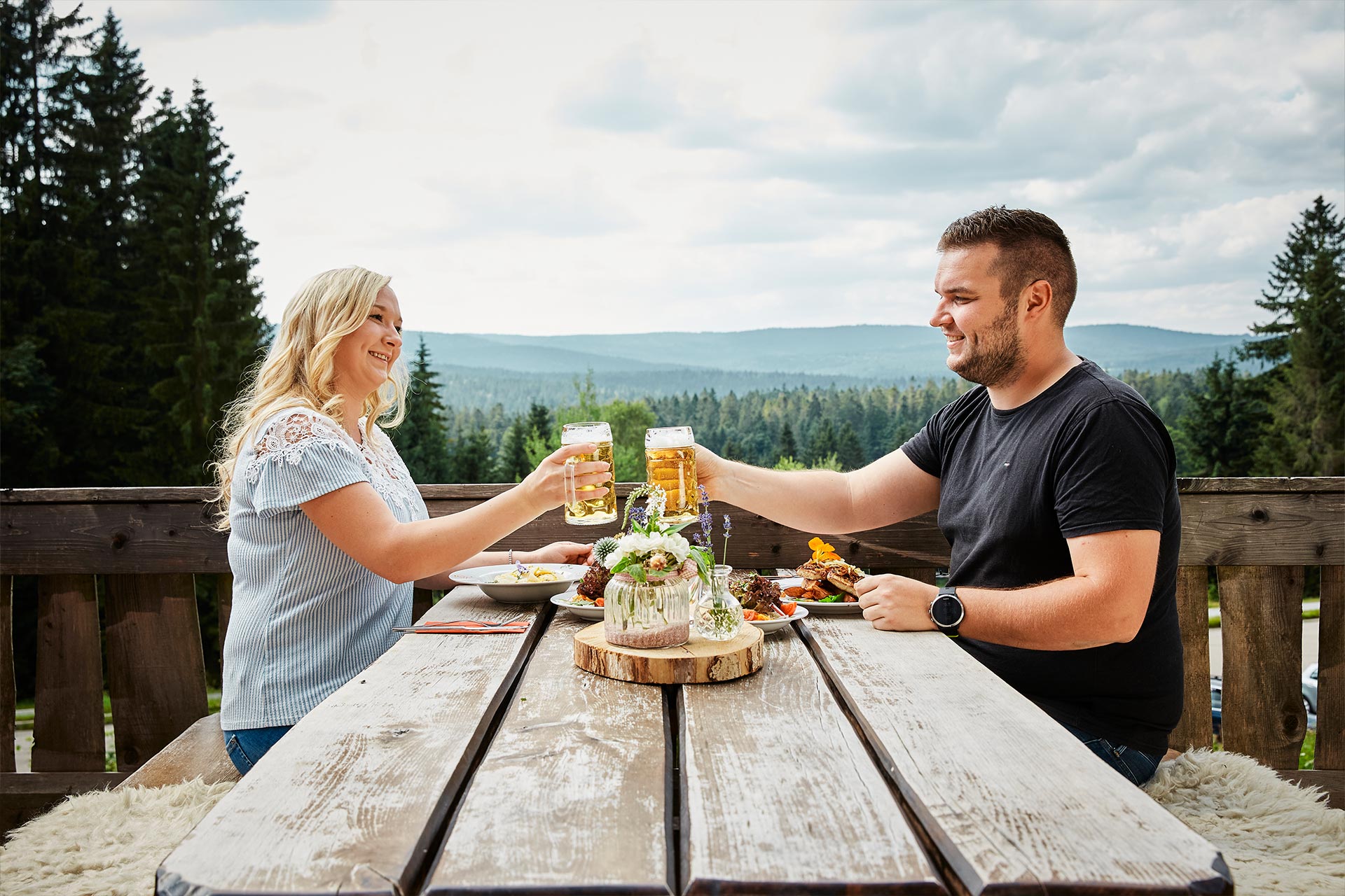 Paar sitzt auf Terrasse eines Gasthauses und stößt mit zwei Bierkrügen an